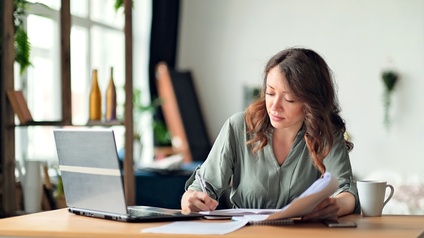 Eine Person sitzt an einem Tisch. Mit der rechten Hand notiert sie etwas in einem Heft. In der linken Hand hält sie Papiere. rechts vor ihr steht ein aufgeklappter Laptop.