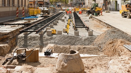 Ein Teil einer Straße ist aufgegraben. In diesem Bereich werden zwei Gleise nebeneinander verlegt. Im Hintergrund stehen Personen sowie verschiedene Bagger. Neben der Straße sind Gebäude