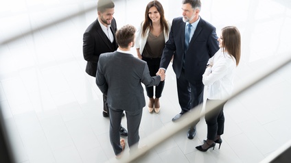 Fünf Personen in Businesskleidung stehen in einem Kreis, zwei der Personen geben sich die Hand