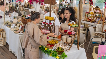 Zwei Personen stehen an einem Marktstand und sprechen miteinander. Auf mehrstöckigen Regalen sind handgefertigte Produkte, darunter Kerzen, Seifen und dekorative Artikel, ausgestellt. Im Hintergrund befinden sich weitere Marktstände sowie mehrere Personen