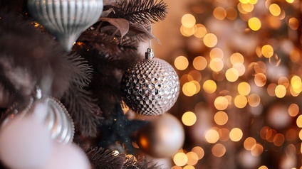 Nahaufnahme einer silbernen Christbaumkugel, die auf einem Zweig eines geschmückten Weihnachtsbaumes hängt, im Hintergrund gelbes und oranges Bokeh