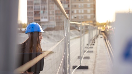 Person mit langen dunklen Haaren und blauem Schutzhelm hält einen Laptop und blickt durch einen Baustellenzaun auf eine Baustelle