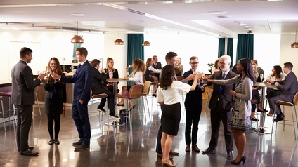 Personen in Businesskleidung stehen und sitzen bei einem Empfang und halten dabei Sektgläser in der Hand