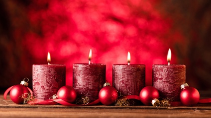 Vier dicke, rote, angezündete Kerzen stehen nebeneinander vor rotem Hintergrund. Vor den Kerzen sind fünf rote, kleine, runde Christbaumkugeln