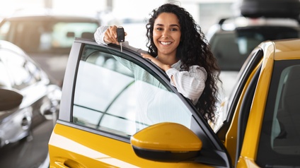 Porträt einer Person mit langen, offenen, dunklen Haaren, die hinter einer geöffneten Autotür steht und den Autoschlüssel hält. Das Auto steht in einer Halle. Daneben stehen weitere Autos