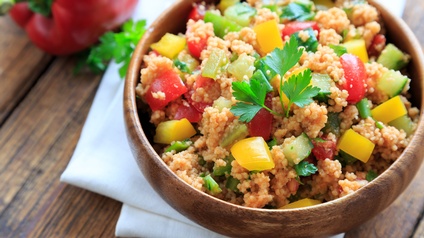 Eine Holzschlüssel mit Couscous, kleinen Gurken und Paprikastückchen sowie Petersilie auf einer weißen Serviette auf braunem Holzuntergrund, daneben liegt eine rote Paprika