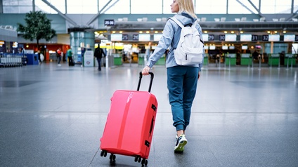Rückenansicht einer Person, die gerade einen Flughafen betritt und ihren roten Koffer mit sich zieht. Am Rücken trägt sie einen weißen Rucksack und schaut auf die Seite.