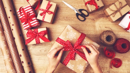 Top-Shot zweier Hände einer Person, die eine rote Masche, die um ein Geschenk drapiert ist, festzieht. Neben dem Geschenk sind weitere Päckchen mit Schliefen, Papierrollen, eine Schere und Geschenkbänder in roten und braunen Farben auf einem Holztisch. 