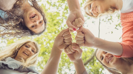 Aufnahme aus der Froschperspektive von vier in einem Kreis stehenden Frauen, die nach unten zur Kamera blicken. In der Bildmitte trifft jeweils eine Faust einer Person aufeinander. Im Hintergrund ist das Geäste eines grünen Baumes