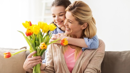 Eine Person sitzt auf einem Sofa und hält in der rechten Hand einen Blumenstrauß aus Tulpen. Die Person wird von hinten von einem Kind umarmt.