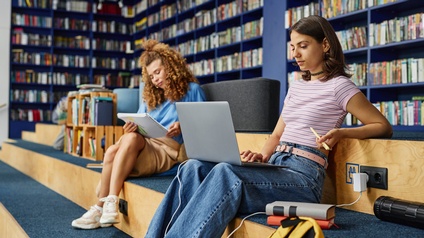 Zwei Personen sitzen auf einer Erhöhung. Die Person links hält ein Notizheft. Die Person rechts hat einen aufgeklappten Laptop am Schoß. Beide blicken auf ihre Gegenstände. An den Wänden stehen mit Büchern befüllte Regale.