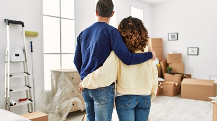 Zwei Personen umarmen sich und blicken in einen Wohnraum mit Umzugskisten, Möbel und Malerutensilien