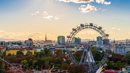 Großes Riesenrad im Wiener Prater mit roten Gondeln im Licht des Sonnenuntergangs, ringsum Baumkronen und im Hintergrund Gebäude der Stadt