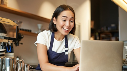 Eine Barista steht hinter dem Tresen und schaut mit einem Lächeln auf den Monitor eines Laptops