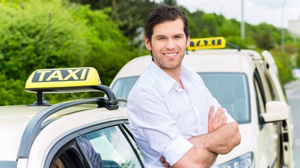 Lächelnde Person mit verschränkten Armen lehnt an Personenkraftwagen mit Taxi-Schild am Autodach, in PKW weitere Person hinter Steuer sitzend, im Hintergrund verschwommen weitere parkende Taxis