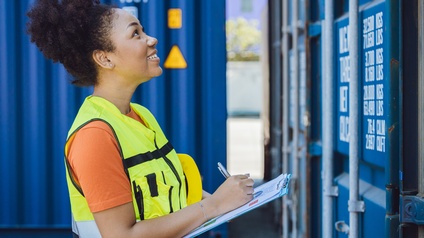 Person mit gelber Warnweste steht fröhlich vor einem Container und notiert sich etwas auf einem Klemmbrett während im Hintergrund weitere blaue Container platziert sind