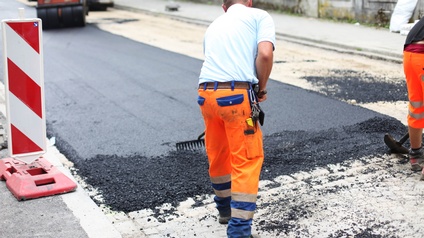 Rückenansicht einer Person, die mit einem Rechen eine teilweise frisch asphaltierte Straße bearbeitet. Im Hintergrund steht ein Fahrzeug mit einer Walze. Links und rechts neben der Straße sind Gehwege
