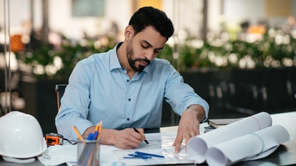 Eine Person sitzt an einem Tisch. Auf dem Tisch liegen eingerollte Papierrollen, Bücher, Stifte, ein Schutzhelm, ein Maßband sowie ein ausgerollter Plan. Die Person hält in der rechten Hand einen Stift.