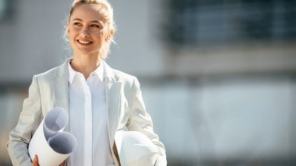 Person in Businesskleidung trägt Papierpläne in einer Hand und mit der anderen Hand einen weißen Schutzhelm 