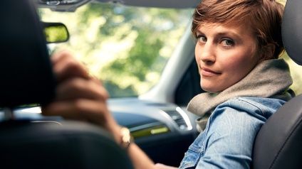 Person mit kurzen braunen Haaren, Schal und Jeansjacke sitzt auf der Beifahrerseite eines Autos und blickt auf die Rückbank zurück in die Kamera