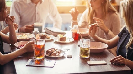 Personen an Tisch sitzend beim Essen. Im Fokus ist ein Holztisch, die Personen sind von der Seite und nur verschwommen zu sehen. Am Tisch sind Stücke von Kuchen und Gläser mit Limonaden platziert. 