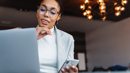Person mit dunklen geschlossenen Haaren, Brille und heller Businesskleidung sitzt in einem Raum mit Luster und blickt gespannt auf den Bildschirm eines Laptops, während sie auch ein Smartphone in der Hand hält