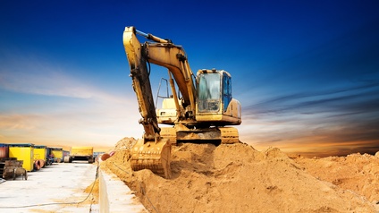 Ein Bagger steht auf einem Erdhaufen neben einer teilweise asphaltieren Straße. Auf der Straße stehen verschiedene Utensilien und weitere Fahrzeuge für den Bau von Straßen