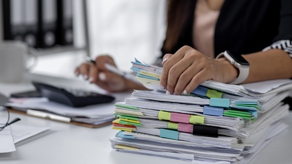 Nahaufnahme eines Stapels Papiere, der auf einem hellen Tisch liegt. Auf dem Stapel liegt die linke Hand einer Person. Die Person sitzt hinter einem Tisch