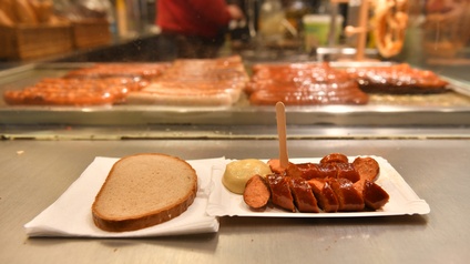 Aufgeschnittene Bratwurst auf Pappteller mit Patzen Senf und Holzgäbelchen in ein Stück gesteckt, nebenliegend auf Serviette Scheibe Brot, im Hintergrund verschwommen weitere Bratwürste auf Bratfläche