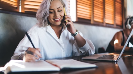 Eine Person sitzt an einem Tisch und telefoniert mit einem Smartphone. Die Person notiert etwas in einem Heft. Daneben ist ein aufgeklappter Laptop. An einem Nebentisch sitzt eine weitere Person