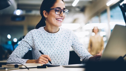 Lächelnde Person mit Brillen sitzt vor Laptop und macht Notizen