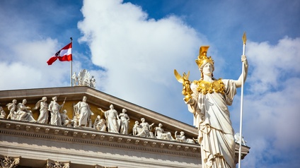 Im Vordergrund ist eine Statue mit goldenem Helm und einem Stab in der Hand. Hinter der Statue ist das Dach eines Hauses aus Stein. Direkt unter dem Dach stehen verschiedene kleinere Statuen. Auf dem Dach ist eine rot-weiß-rote Flagge