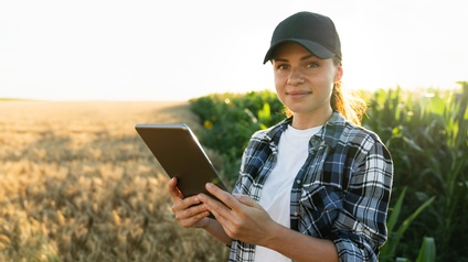 Portrait einer sanft lächelnden Person im Licht des Sonnenuntergangs mit Kappe, die Tablet in Händen hält und auf Feld neben Maispflanzen steht