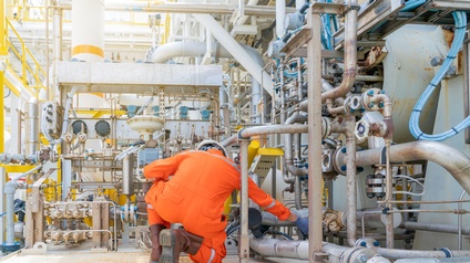 Person in orangem Schutzanzug mit Helm in Rückenansicht hockt am Boden vor großem Rohrsystem in einer Raffinerie und greift nach einer Leitung