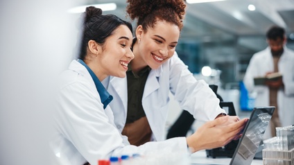 Zwei Personen in weißen Kitteln stehen nebeneinander an einem Tisch. Vor ihnen steht ein aufgeklappter Laptop, auf den beide blicken. Daneben sind mehrere Reagenzgläser. Im Hintergrund ist eine weitere Person in einem weißen Kittel