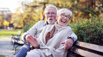 Paar mittleren Alters mit Brille sitzen freudig auf einer Parkbank im Herbst
