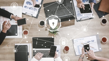 Top-Shot einer hölzernen Oberfläche, auf der aufgeklappte Laptops, Klemmbretter mit Papieren mit Statistiken, Tassen mit Flüssigkeiten, die Hände von Personen mit Smartphones sind. Darüber sind kleine Schlösser umgeben von Kreisen