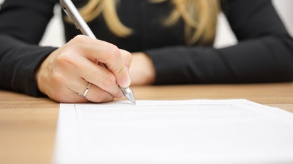 Eine Person unterschreibt mit einem Kugelschreiber auf einem bedruckten Blatt Papier