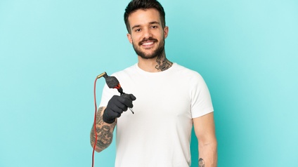 Porträt einer lächelnden Person mit dunklen kurzen Haaren und Bart hält eine Tätowiermaschine