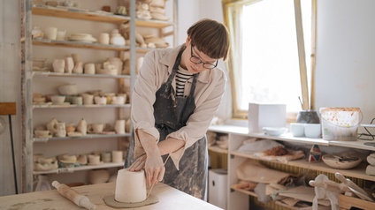 Person mit Brillen in Schürze steht an Tisch in Töpfereiwerkstatt und bearbeitet Ton mit Holzstäbchen, im Hintergrund Regale mit Töpferwaren