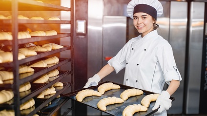 Person in weißem Kochgewand mit Kochhaube hält Blech mit Croissants in Händen