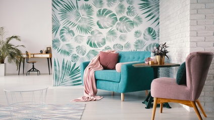 Türkises Sofa mit Kissen und Decke vor tapezierter Wand mit Blattmuster, ringsum weitere Einrichtungsgegenstände