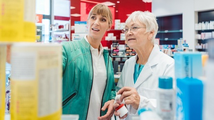 Frontalaufnahme zweier nebeneinander stehender Personen. Die rechte Person trägt einen weißen Kittel. Beide blicken in die linke Bildhälfte. Im Hintergrund sind Regale, befüllt mit Objekten. Im Vordergrund sind Verpackungen und Tuben