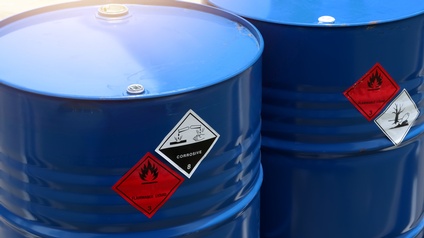 Zwei blaue Metallfässer mit roten quadratischen Stickern: Schwarzes Symbol einer Flamme, Schriftzug Flammable Liquid 3 und weiße quadratische Sticker mit Phiolen, die Flüssigkeit auf Hand & Baum tropfen sowie Fisch & blattloser Baum