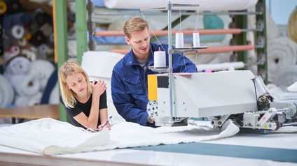 Zwei Personen stehen bei einer Industrie-Nähmaschine, eine Person mit Bart trägt eine blaue Arbeitsbekleidung, im Hintergrund sind mehrere Rollen Stoff bereit zur Verarbeitung auf Rollen oder in Regalen geschlichtet