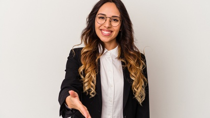 Porträt einer lächelnden Person mit offenen, lockigen Haaren, Brille, weißer Bluse und dunklem Sakko, die ihre rechte Hand in Richtung Kamera streckt