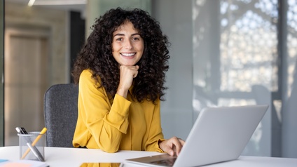 Porträt einer lächelnden Person mit dunklen, offenen, lockigen Haaren und gelblicher Bluse, die an einem Tisch sitzt und vor sich einen Laptop, ein Tablet und Stifte hat