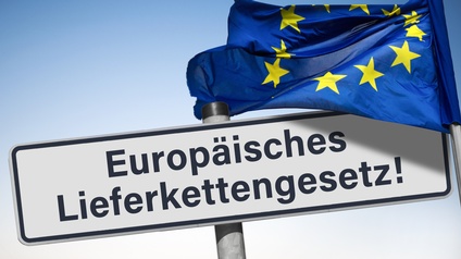 Auf einem weißen Schild steht europäisches Lieferkettengesetz. Rechts daneben ist eine blaue Flagge mit gelben Sternen in einem Kreis