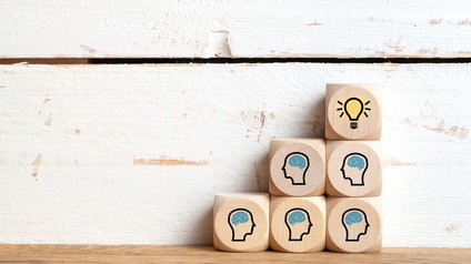 Vor einer hellen Wand stehen fünf Holzwürfel übereinander. Auf fünf Würfeln ist der Umriss eines menschlichen Kopfes, in dem jeweils das Gehirn blau eingezeichnet ist. Auf dem sechsten und obersten Würfel ist eine leuchtende Glühbirne