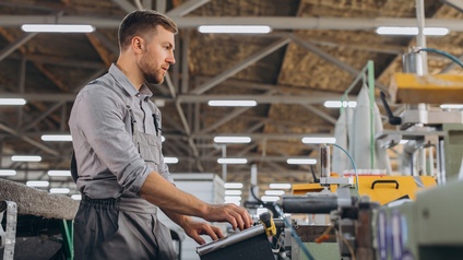 Seitliche Aufnahme einer stehenden Person, die ihre Hände auf einer metallenen Vorrichtung hat, mit der er eine Maschine bewegen kann, in einer großen Halle mit Deckenlampen.